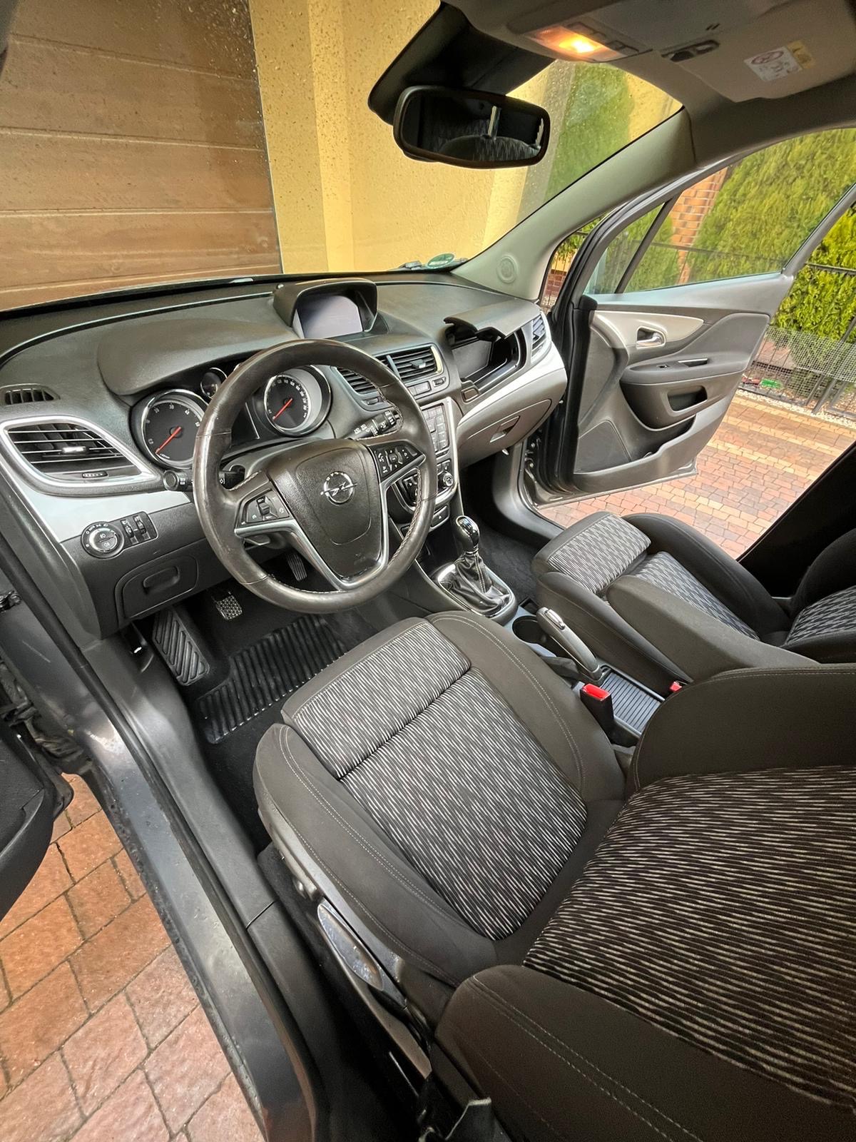 Wnętrze samochodu Opel Mokka z widocznymi fotelami z szaro-białą tapicerką w paski, deską rozdzielczą i kierownicą z logo marki. Widoczne otwarte drzwi od strony kierowcy.