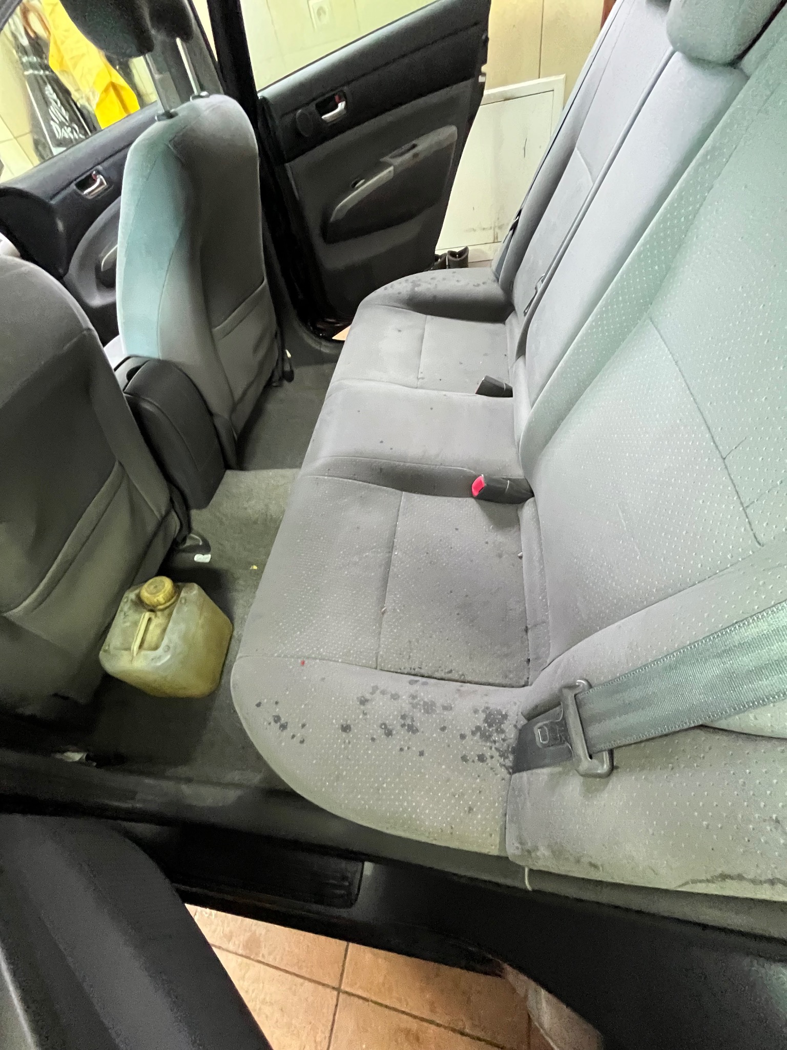 Wnętrze samochodu z widocznymi, mocno zabrudzonymi tylnymi siedzeniami tapicerowanymi, obok których stoi plastikowy kanister. Widoczne plamy i zacieki na tapicerce.