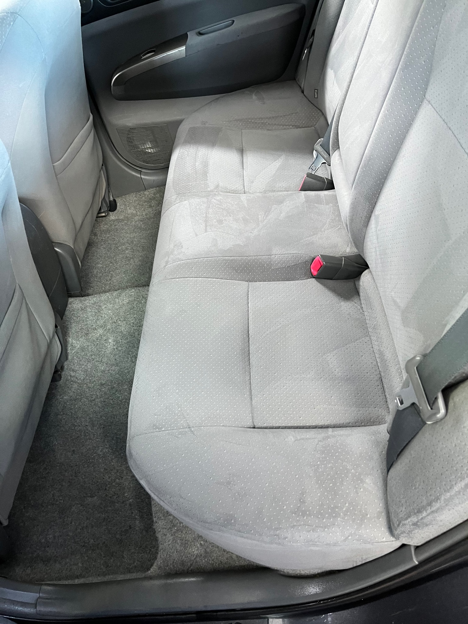 Wnętrze samochodu z szarą tapicerką po czyszczeniu, widoczne wilgotne plamy na tylnej kanapie i dywanikach, zapięty pas bezpieczeństwa z różowym elementem.
