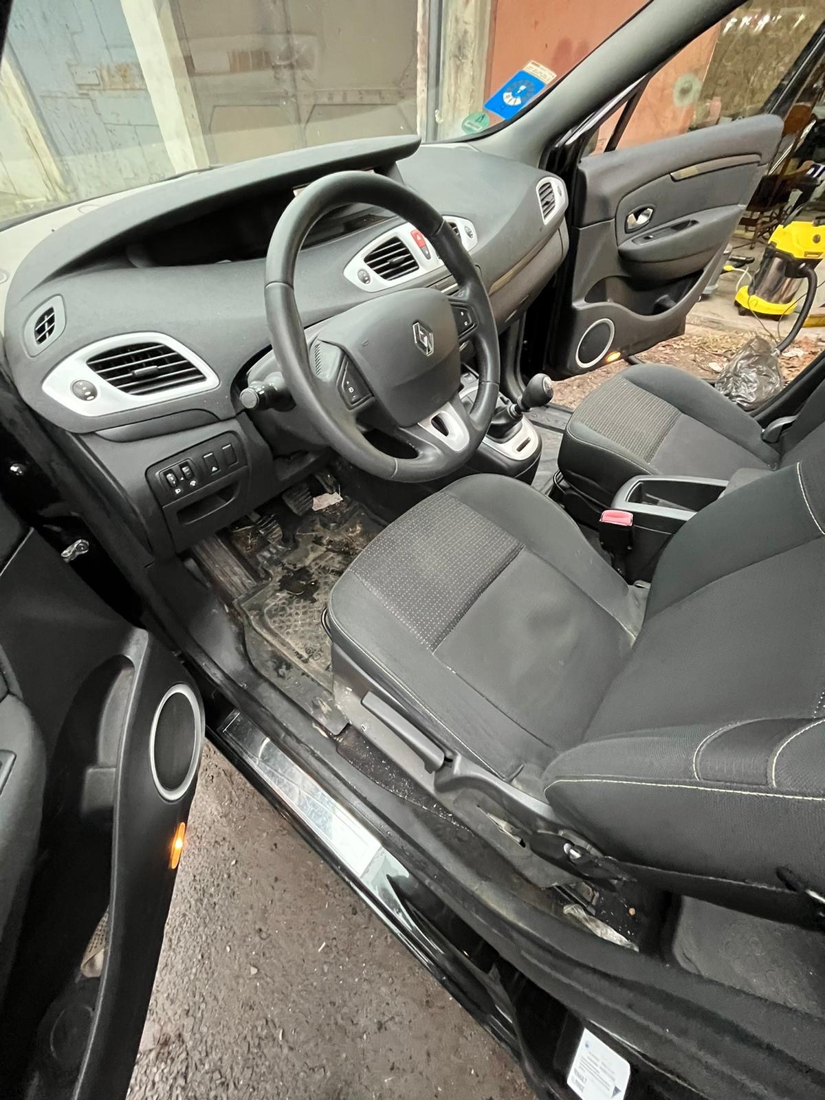 Wnętrze czarnego samochodu Renault Scenic przed czyszczeniem: brudne dywaniki, tapicerka foteli z plamami, widoczny odkurzacz piorący w tle.