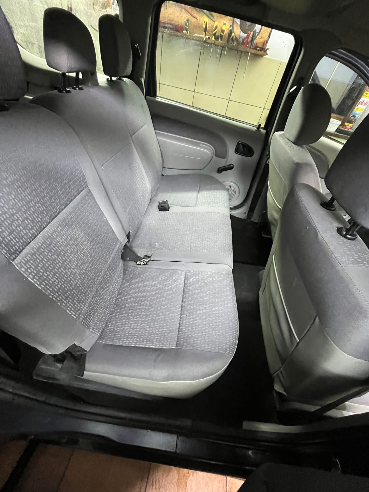 Wnętrze samochodu z widocznymi szarymi, tekstylnymi siedzeniami tylnymi z pasami bezpieczeństwa, widoczne narzędzia na ścianie w tle.