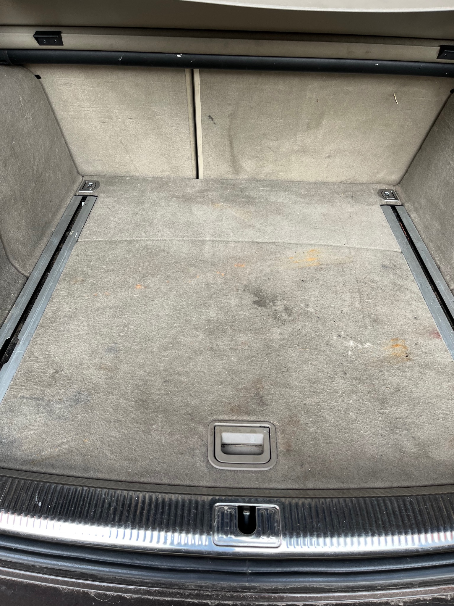 Wnętrze bagażnika samochodowego z widocznymi plamami na tapicerce podłogi i metalowymi prowadnicami.