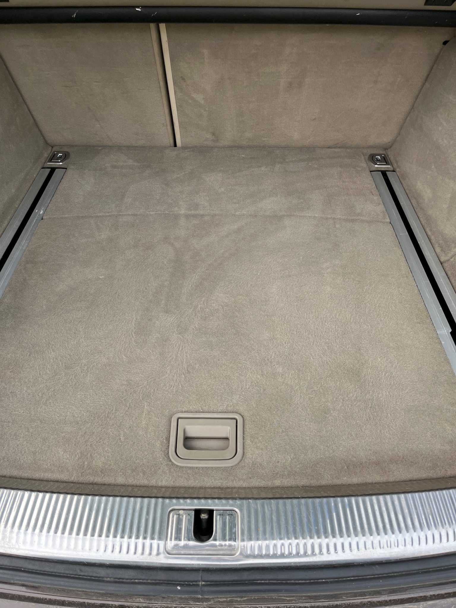 Wnętrze bagażnika samochodu z beżową tapicerką, widoczne metalowe szyny i uchwyt do podnoszenia podłogi.