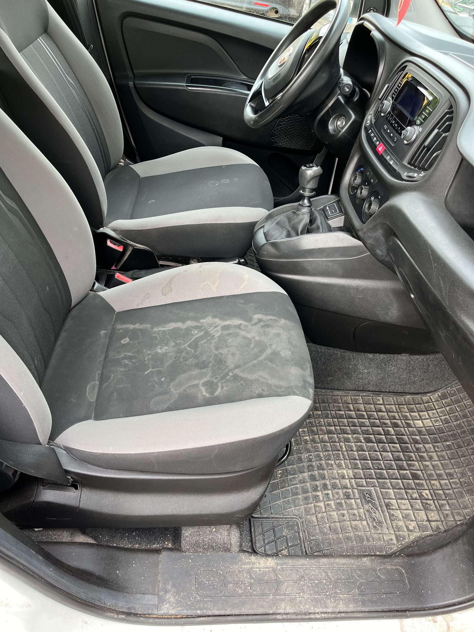 Brudne, szaro-czarne fotele tapicerowane w samochodzie, widoczne plamy i zabrudzenia, gumowa wycieraczka pod nogami, wnętrze pojazdu w Legnicy przed czyszczeniem.