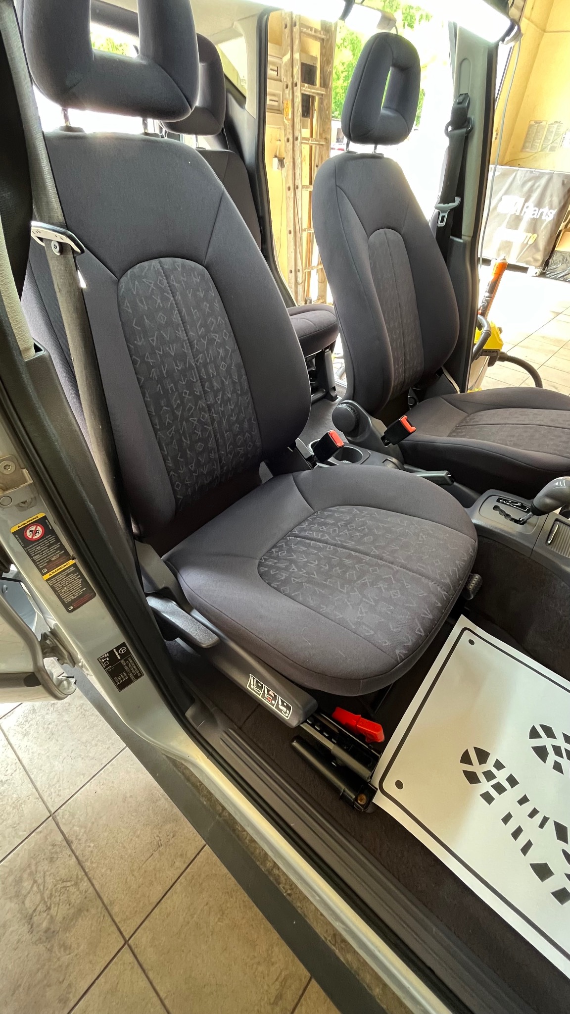 Wnętrze samochodu z dwoma czarnymi, tapicerowanymi fotelami z wzorem, widoczne z otwartymi drzwiami, z naklejką z symbolem butów na podłodze.