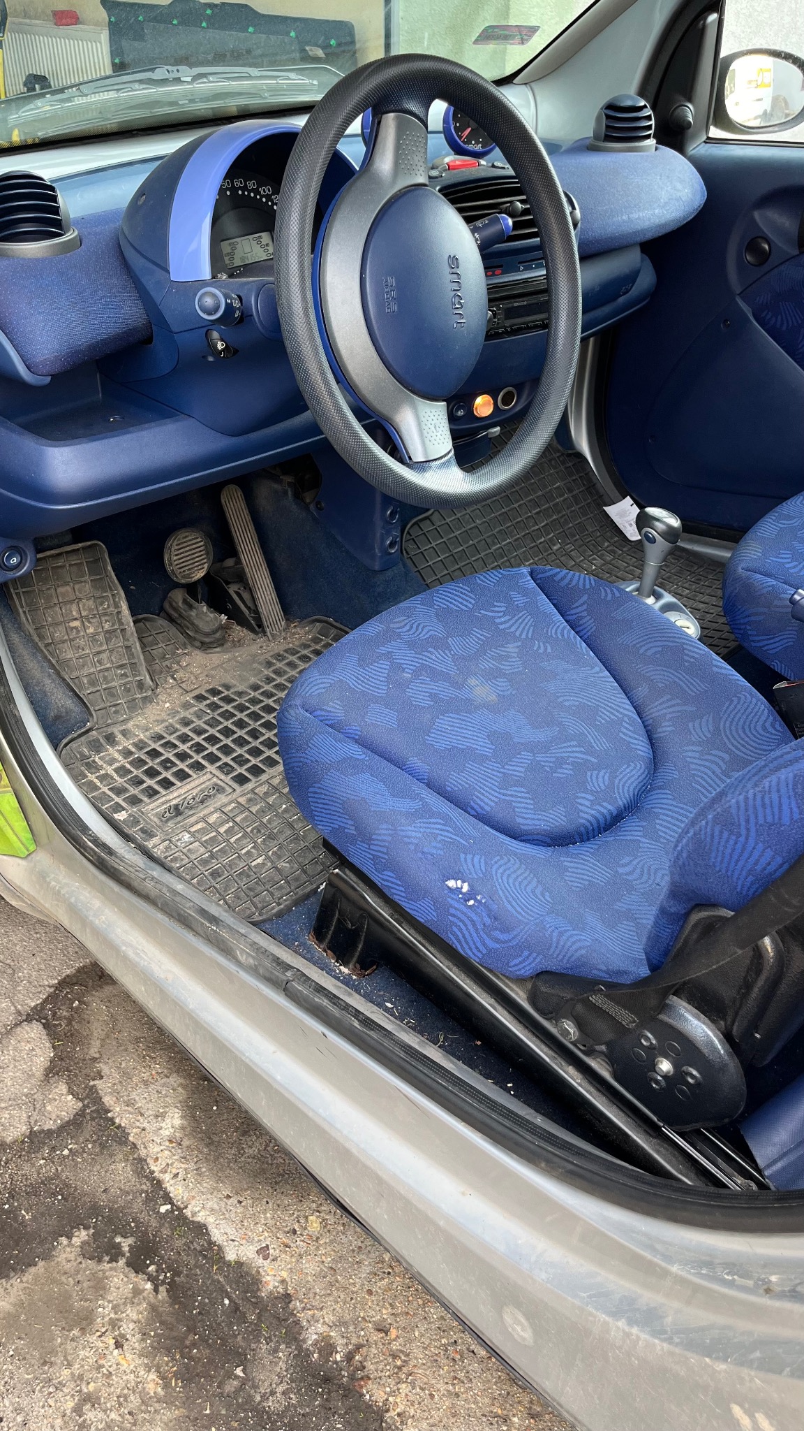 Wnętrze samochodu Smart z niebieską tapicerką, widoczne zabrudzenia na dywanikach i plamy na siedzeniu kierowcy.