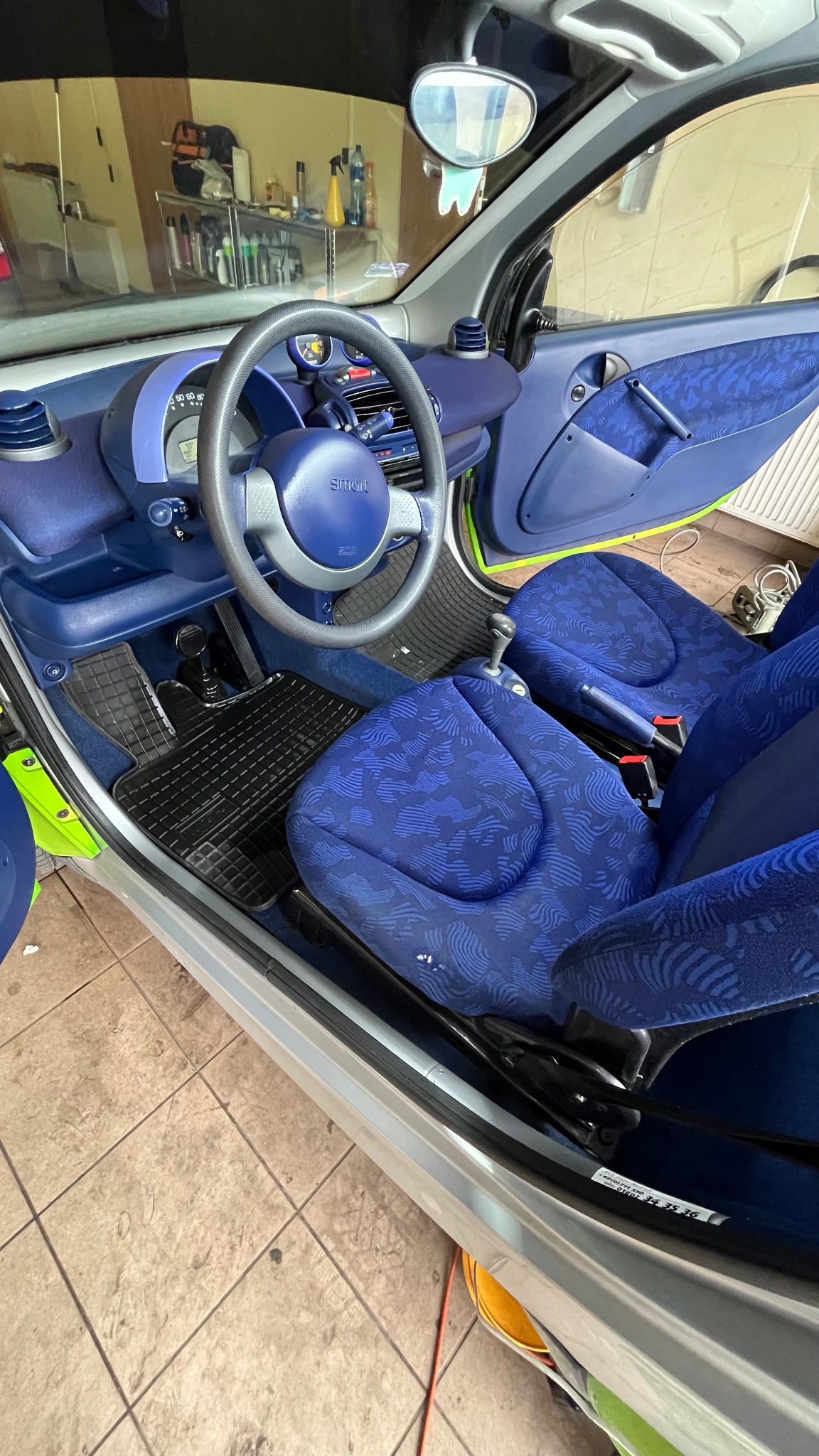 Wnętrze samochodu Smart z niebieską tapicerką w geometryczne wzory, widoczne fotele, kierownica i deska rozdzielcza, otwarte drzwi po stronie kierowcy.