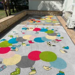 Dwa dywany dziecięce z kolorowymi wzorami na kostce brukowej przed budynkiem, jeden z motywem gąsienicy z kół, drugi z motylami i kwiatami.