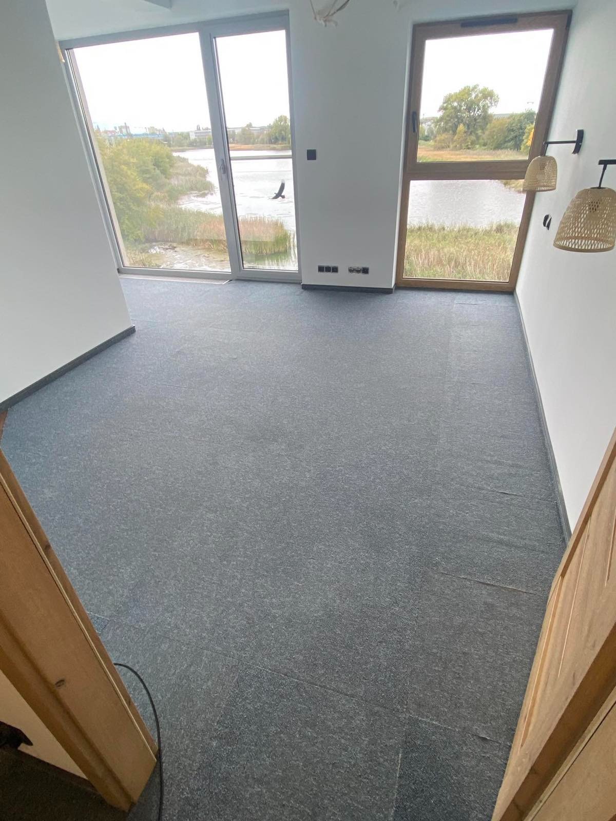 Wnętrze pokoju z widokiem na jezioro przez duże okna, szara wykładzina podłogowa, drewniane ramy okienne i drzwi, oraz dekoracyjne lampy ścienne.