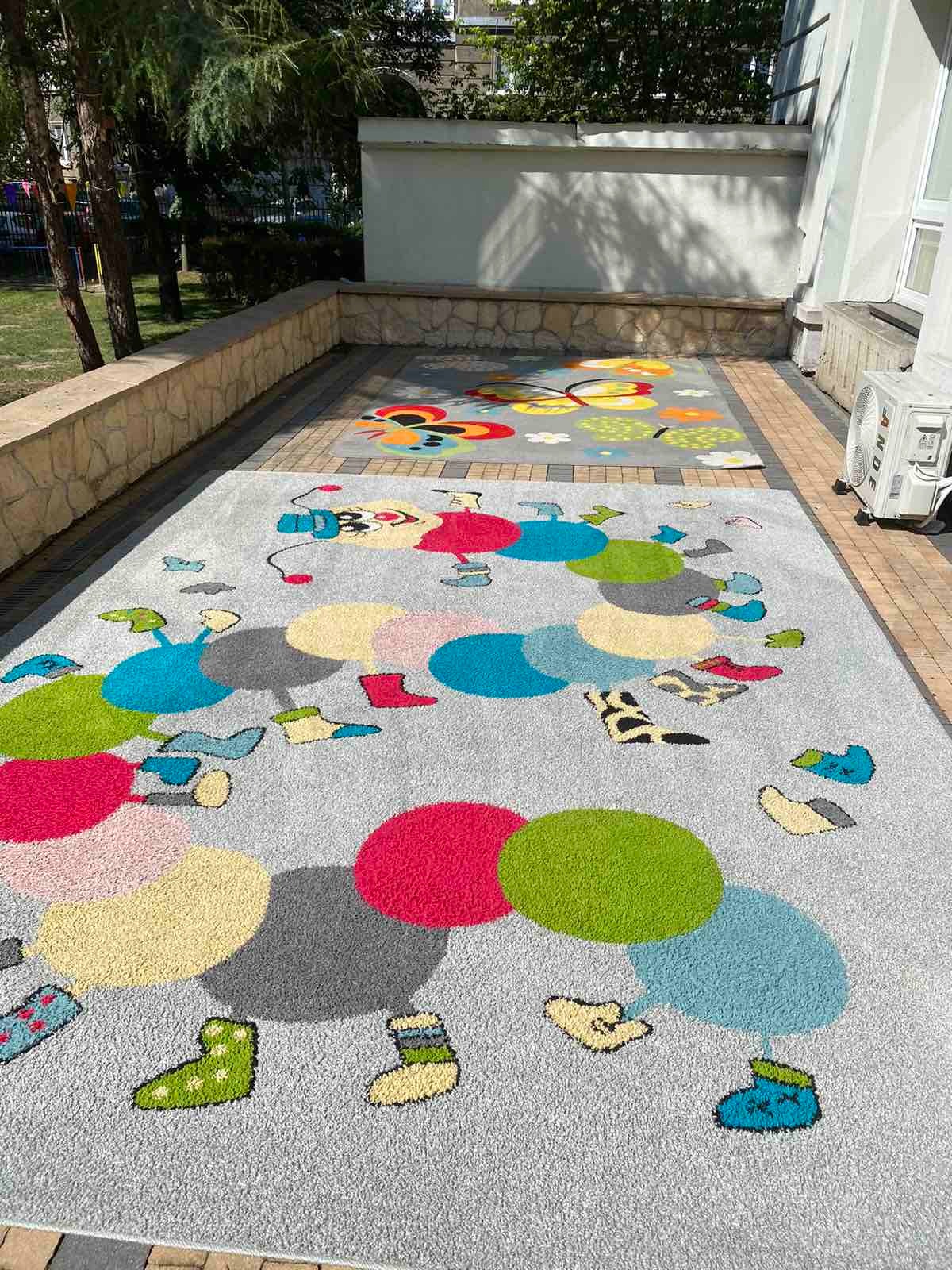 Dwa dywany dziecięce z kolorowymi wzorami na kostce brukowej przed budynkiem, jeden z motywem gąsienicy z kół, drugi z motylami i kwiatami.