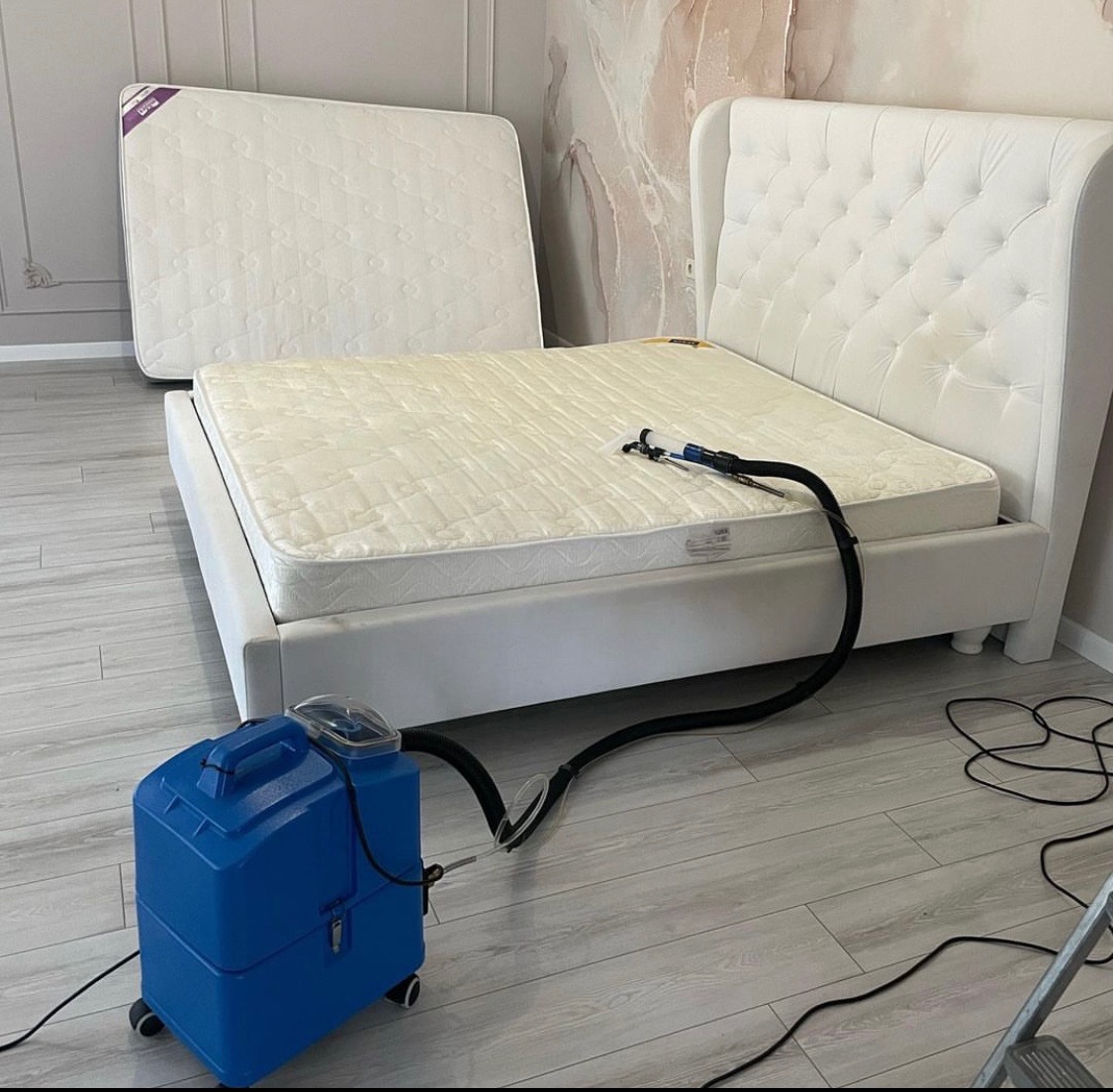 Białe łóżko z materacem poddawane czyszczeniu parowemu niebieskim odkurzaczem piorącym; drugi materac oparty o ścianę w tle.