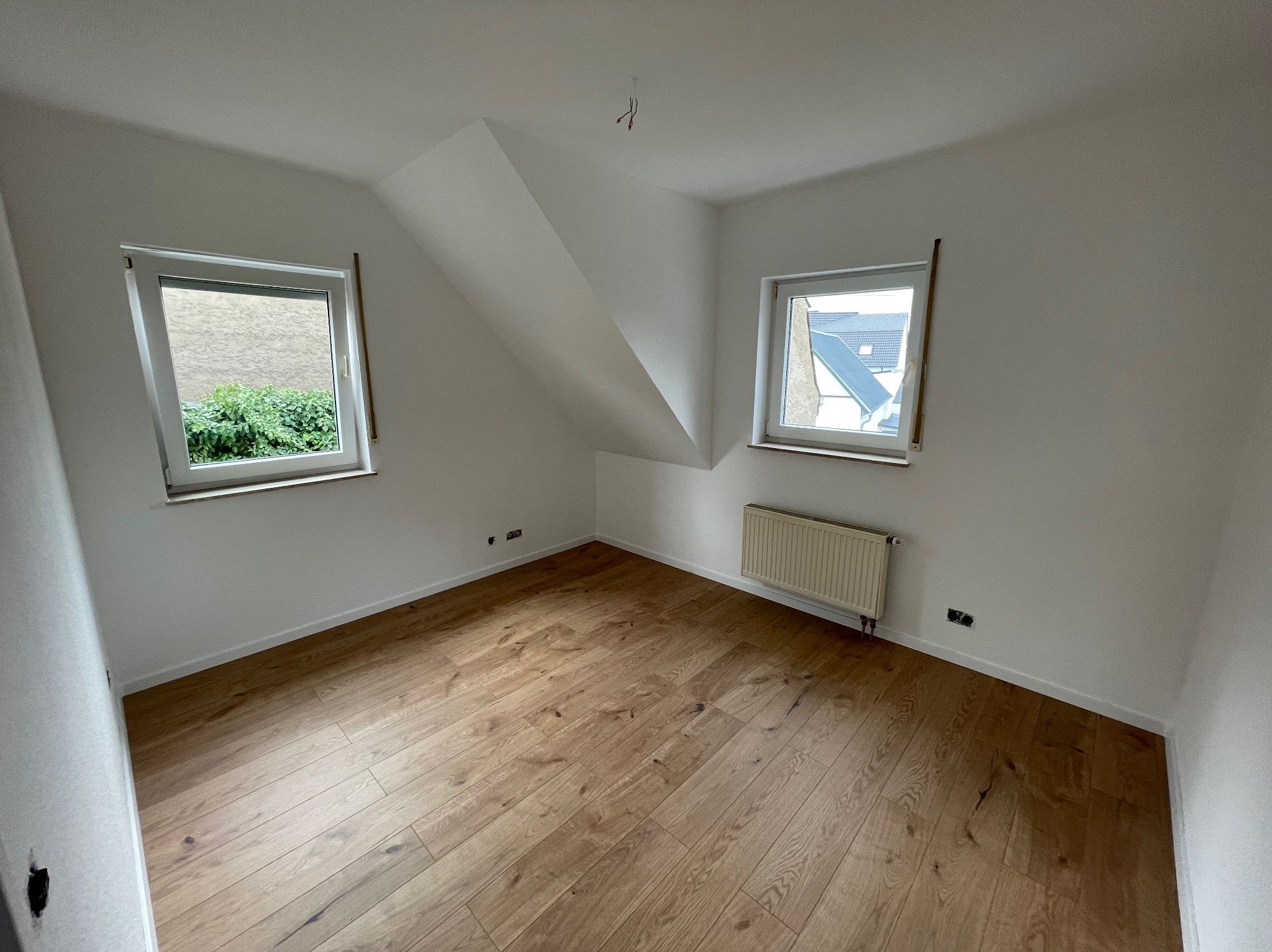 Puste poddasze z dwiema oknami, drewnianą podłogą i grzejnikiem, świeżo po remoncie, z widocznymi przewodami elektrycznymi na suficie.