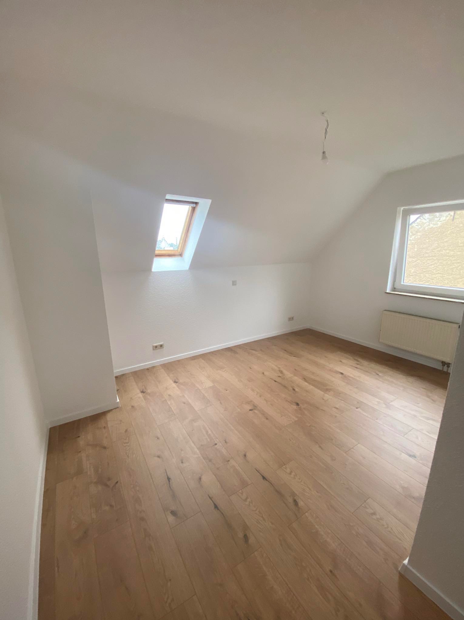 Poddasze po remoncie: drewniana podłoga, białe ściany, okno dachowe i grzejnik pod oknem na ścianie szczytowej, widoczne gniazdka elektryczne i wystający kabel po lampie.