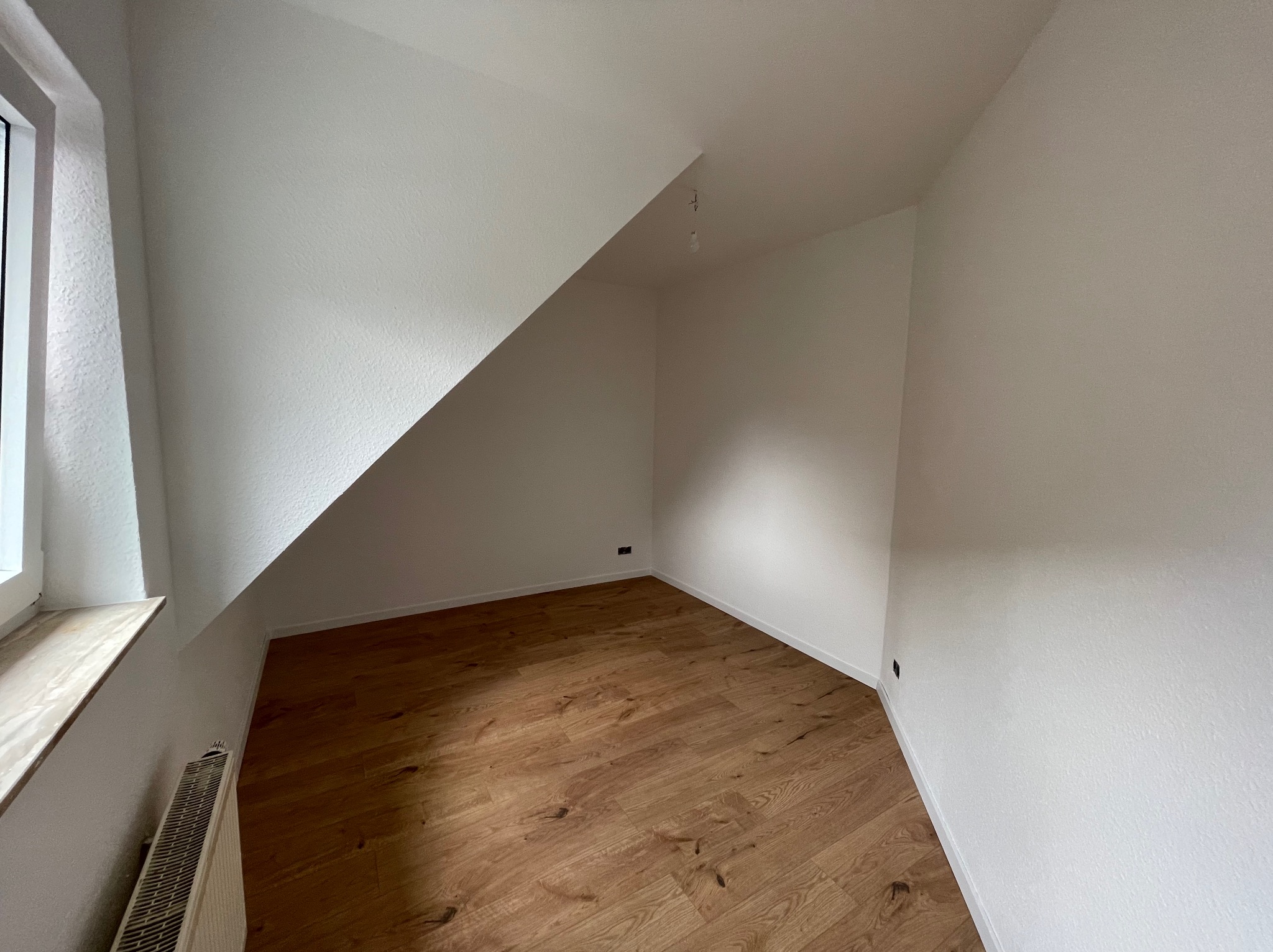Puste, świeżo wyremontowane poddasze ze skośnym sufitem, drewnianą podłogą i białymi ścianami, widoczne gniazdka elektryczne, brak lampy.