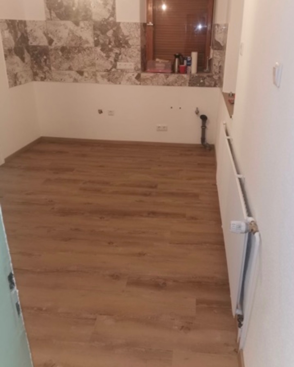 Pusty pokój po remoncie z nową drewnianą podłogą, częściowo wykończoną ścianą z brązowych płytek i grzejnikiem przy ścianie.