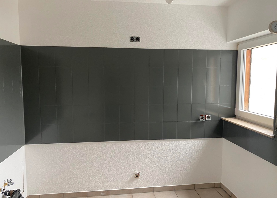 Świeżo położone, błyszczące, grafitowe płytki ścienne w kuchni, z widocznymi puszkami elektrycznymi i fragmentem okna, dolna część ściany pomalowana na biało, jasna podłoga z płytek.