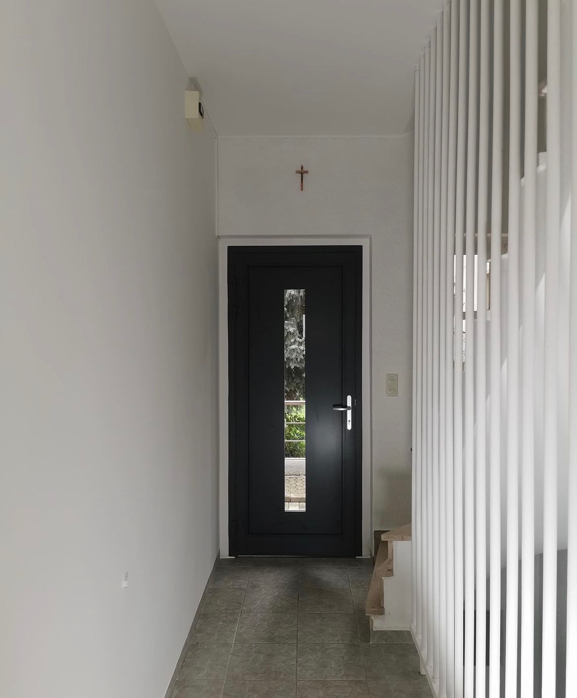 Wnętrze korytarza z szarą podłogą, czarnymi drzwiami z przeszkleniem oraz białą ścianą z drewnianym krzyżem i pionowymi białymi listwami przy schodach.