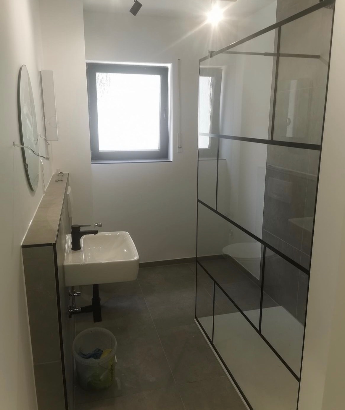 Nowoczesna łazienka po remoncie z białą umywalką nablatową z czarną armaturą, szarą podłogą i kabiną prysznicową z czarnymi profilami oraz widocznym wiadrem z akcesoriami do sprzątania.