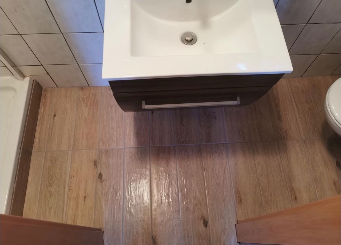 Ujęcie z góry na umywalkę z szafką w brązowym odcieniu drewna, jasną podłogę imitującą drewno i fragment toalety oraz kabiny prysznicowej w tle, ukazujące wykończenie łazienki po remoncie.