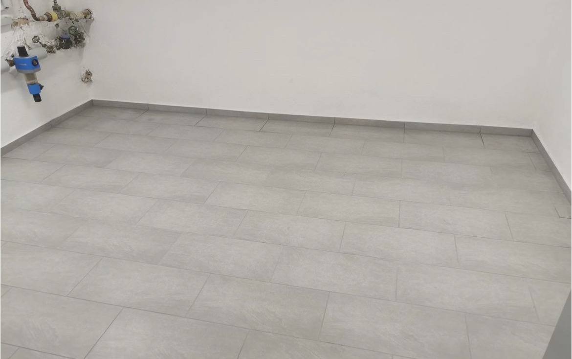 Wykończone wnętrze z szarymi, prostokątnymi płytkami podłogowymi, widoczne rury i instalacje przy ścianie w górnym lewym rogu.