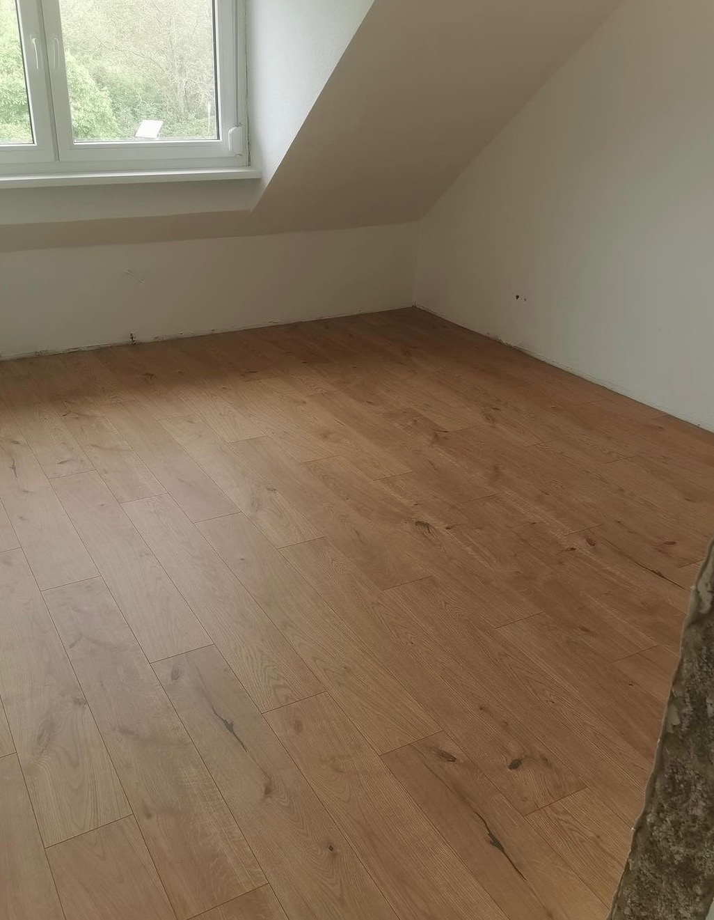 Jasne panele podłogowe ułożone w pokoju na poddaszu z oknem, widoczne ślady po remoncie na ścianach.