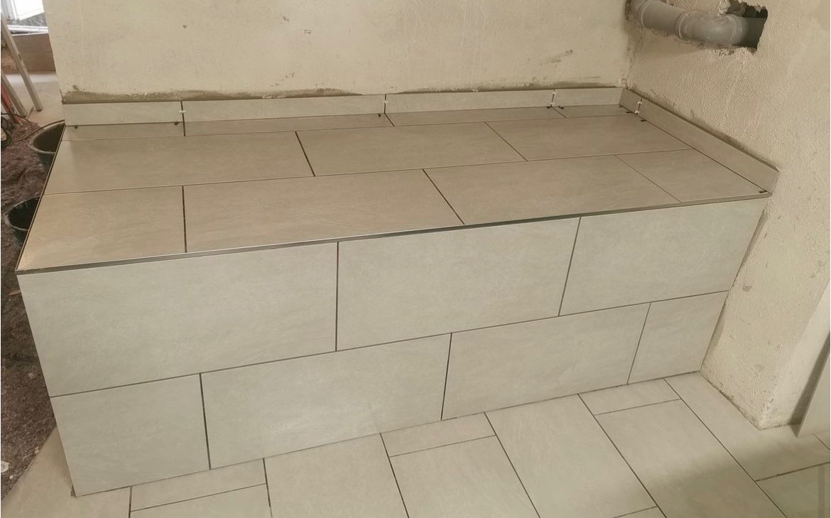 Wykończona obudowa wanny lub brodzika z jasnych, prostokątnych płytek ceramicznych z metalową listwą wykończeniową, widoczna rura kanalizacyjna w górnym rogu, surowa ściana w tle.