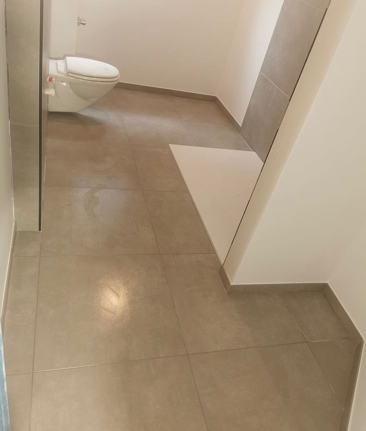 Wykończona łazienka z beżowymi płytkami podłogowymi i częściowo wykończoną ścianą, widoczny biały sedes podwieszany.