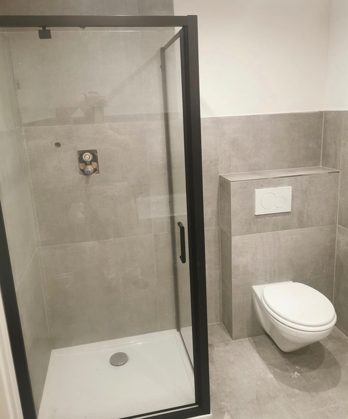 Wykończona łazienka z kabiną prysznicową z czarną ramą, wiszącą toaletą i szarą okładziną ścienną imitującą beton.