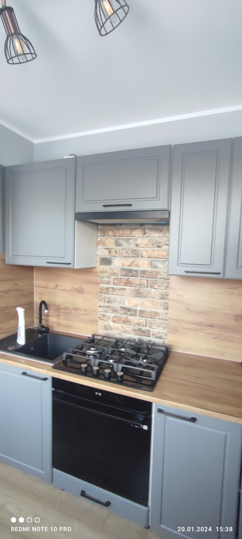 Szare szafki kuchenne z czarnym zlewem i płytą gazową, wykończone drewnopodobnym blatem i ceglaną ścianą nad płytą.