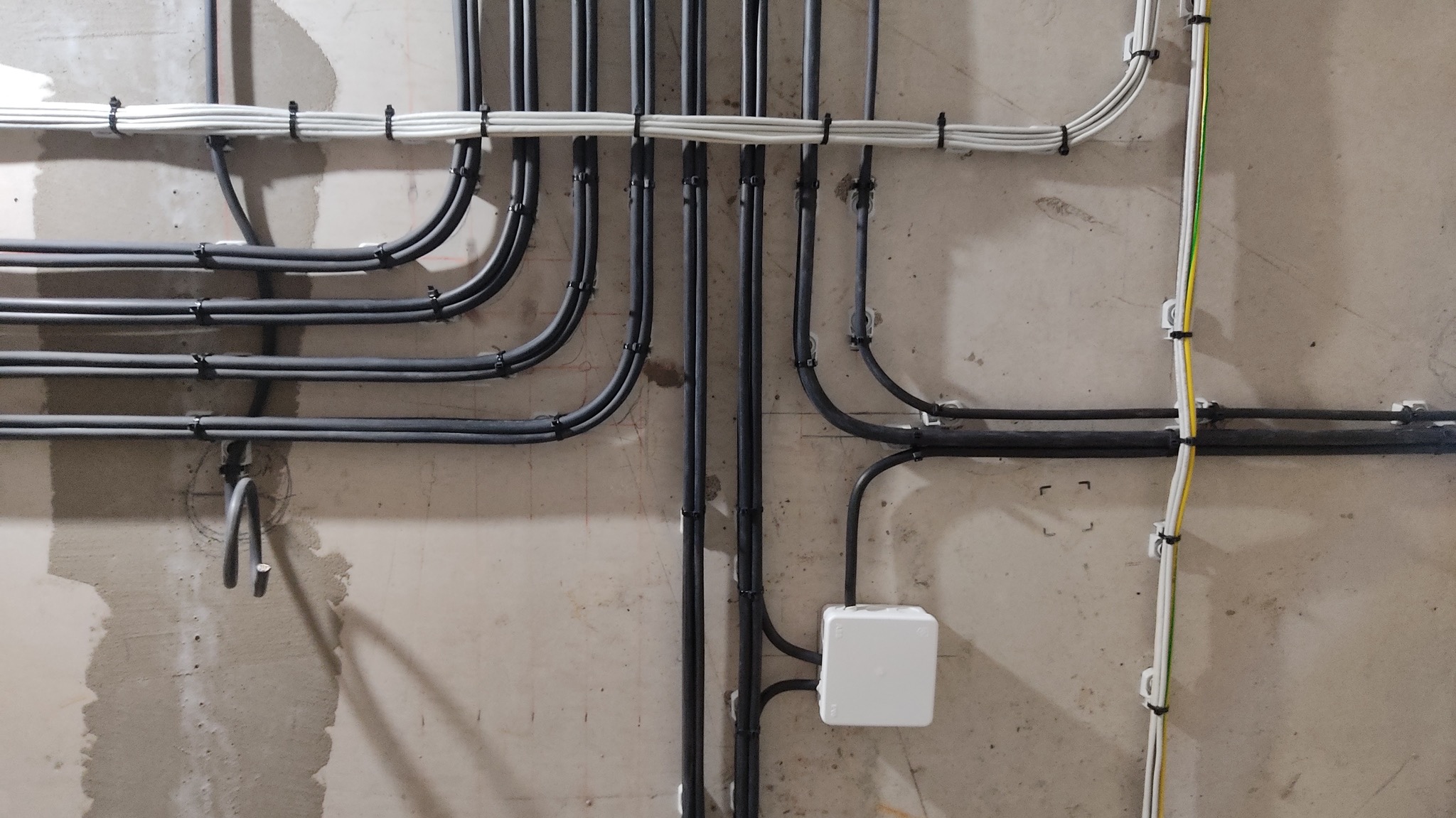 Szare i białe kable elektryczne starannie ułożone na surowej ścianie, zabezpieczone uchwytami. Widoczna puszka instalacyjna.