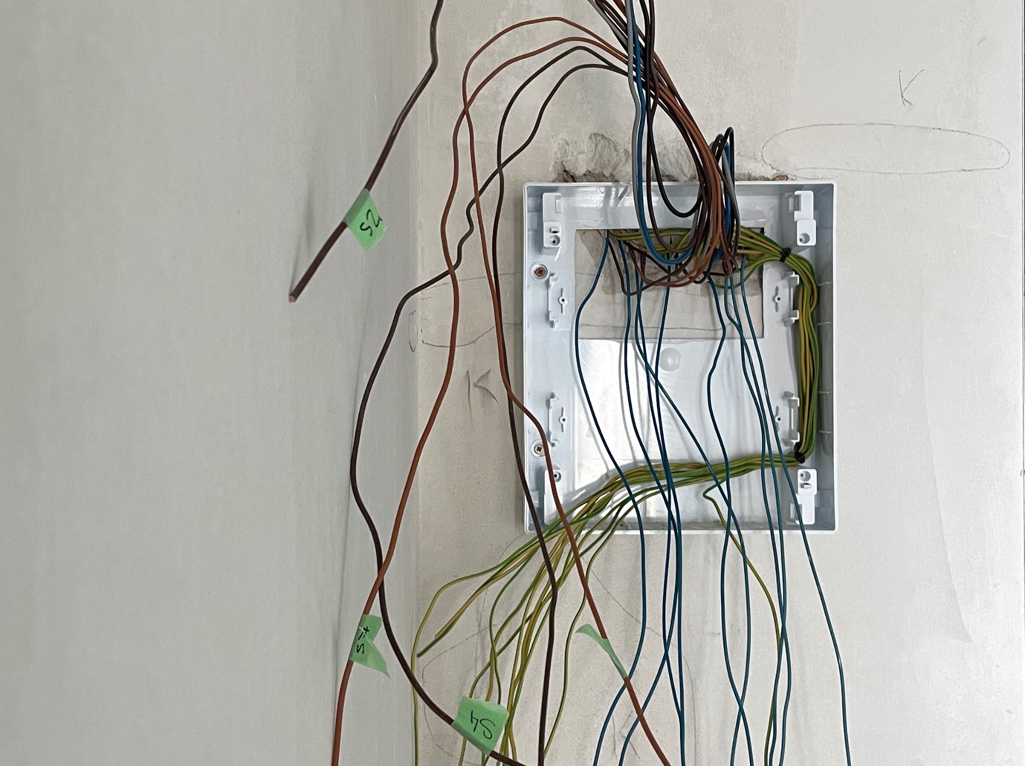 Biała puszka elektryczna na ścianie z wystającymi przewodami oznaczonymi zielonymi etykietami, widoczne ślady po wierceniu.