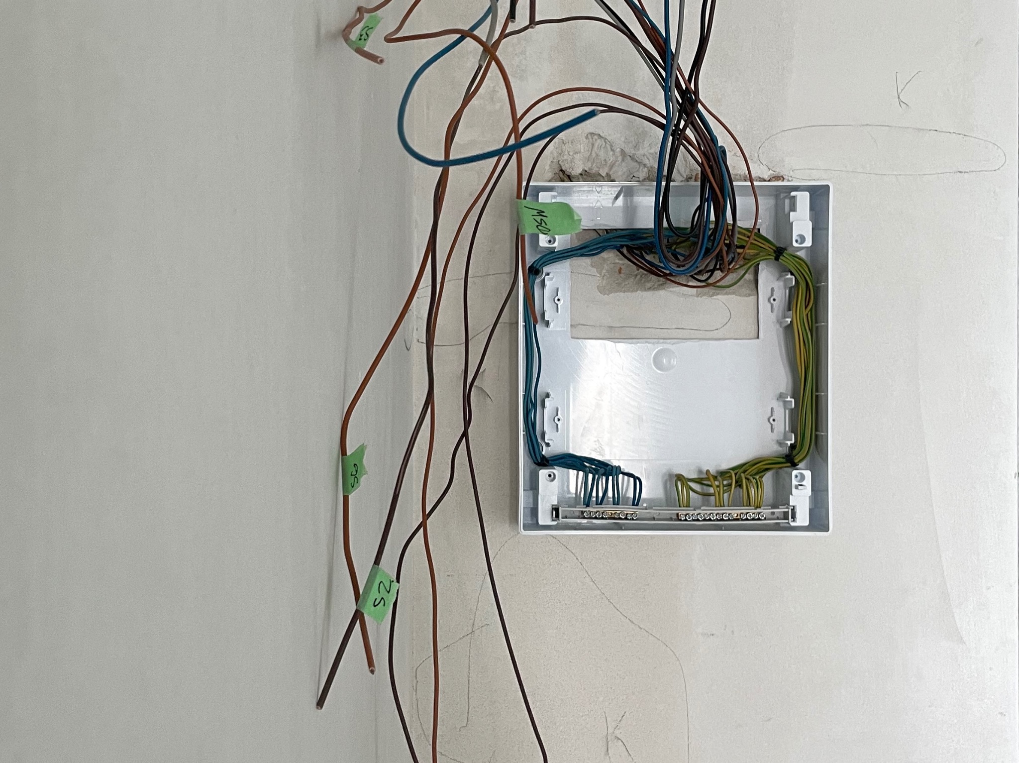 Otwarta, biała puszka elektryczna w ścianie z wystającymi kablami oznaczonymi zielonymi etykietami. Widoczne przewody wewnątrz i na zewnątrz puszki. Etap prac instalacyjnych.