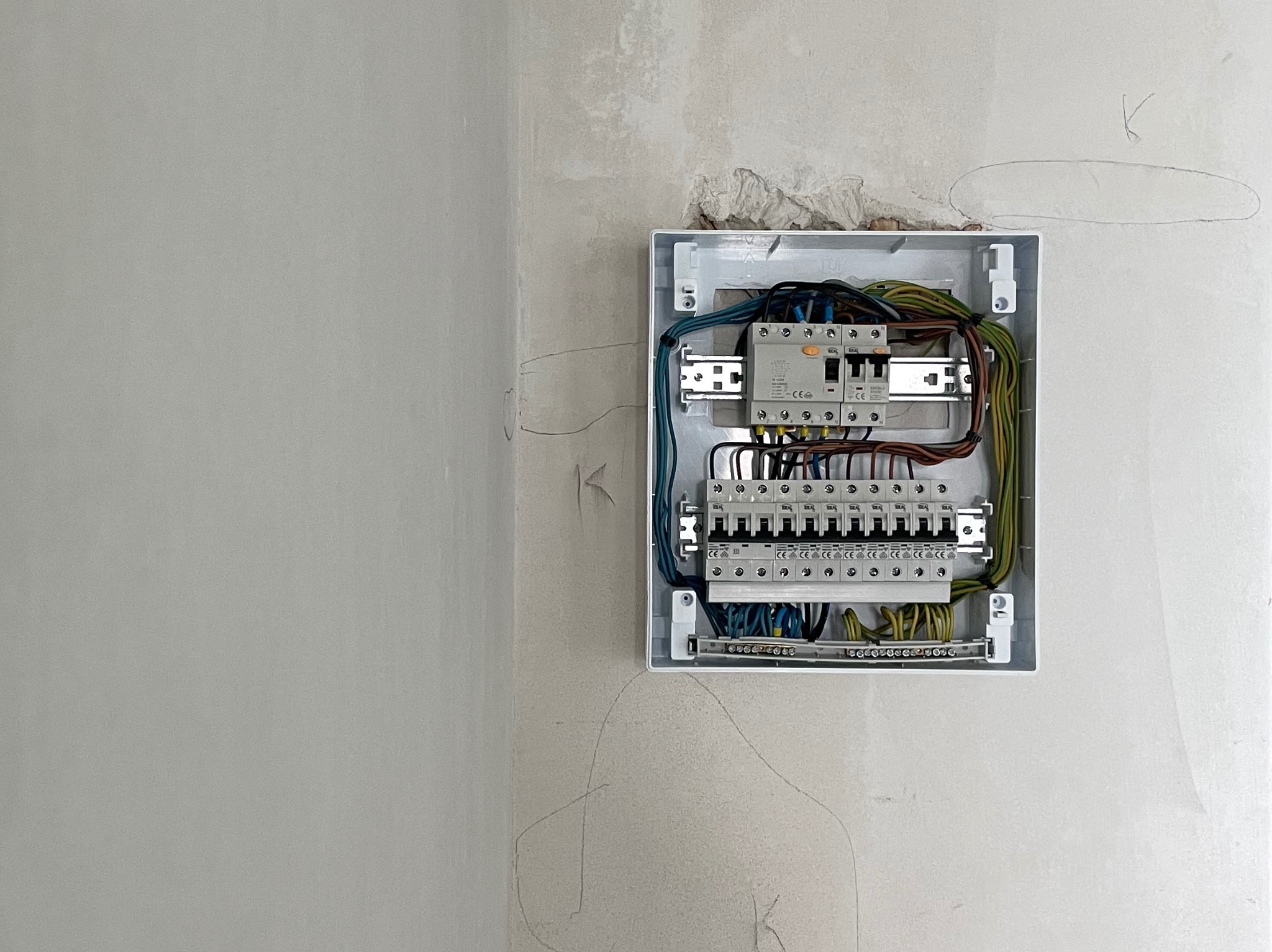 Rozdzielnica elektryczna w białej obudowie, zamontowana na szarej ścianie w trakcie prac wykończeniowych. Widoczne kable i zabezpieczenia.