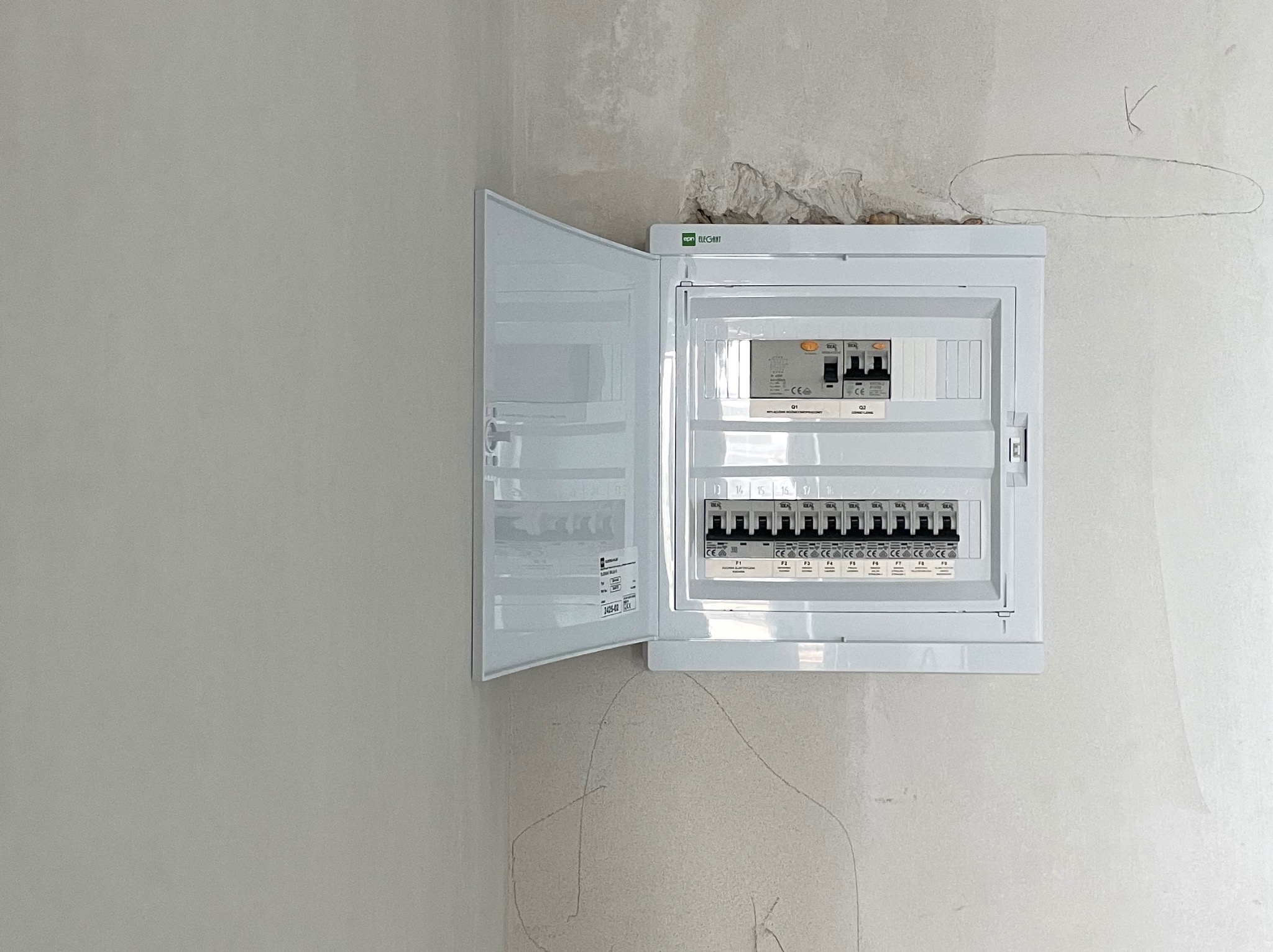 Biała skrzynka rozdzielcza z otwartymi drzwiczkami, zamontowana na surowej ścianie, widoczne bezpieczniki i oznaczenia.