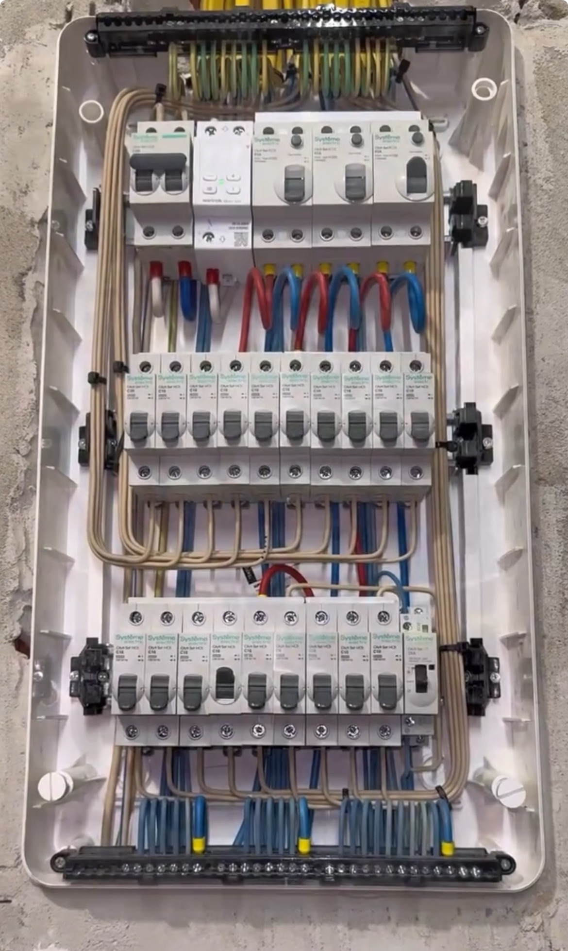 Rozdzielnica elektryczna w białej obudowie, z widocznymi bezpiecznikami, przewodami w kolorach niebieskim i czerwonym, zamontowana na szarej, surowej ścianie.