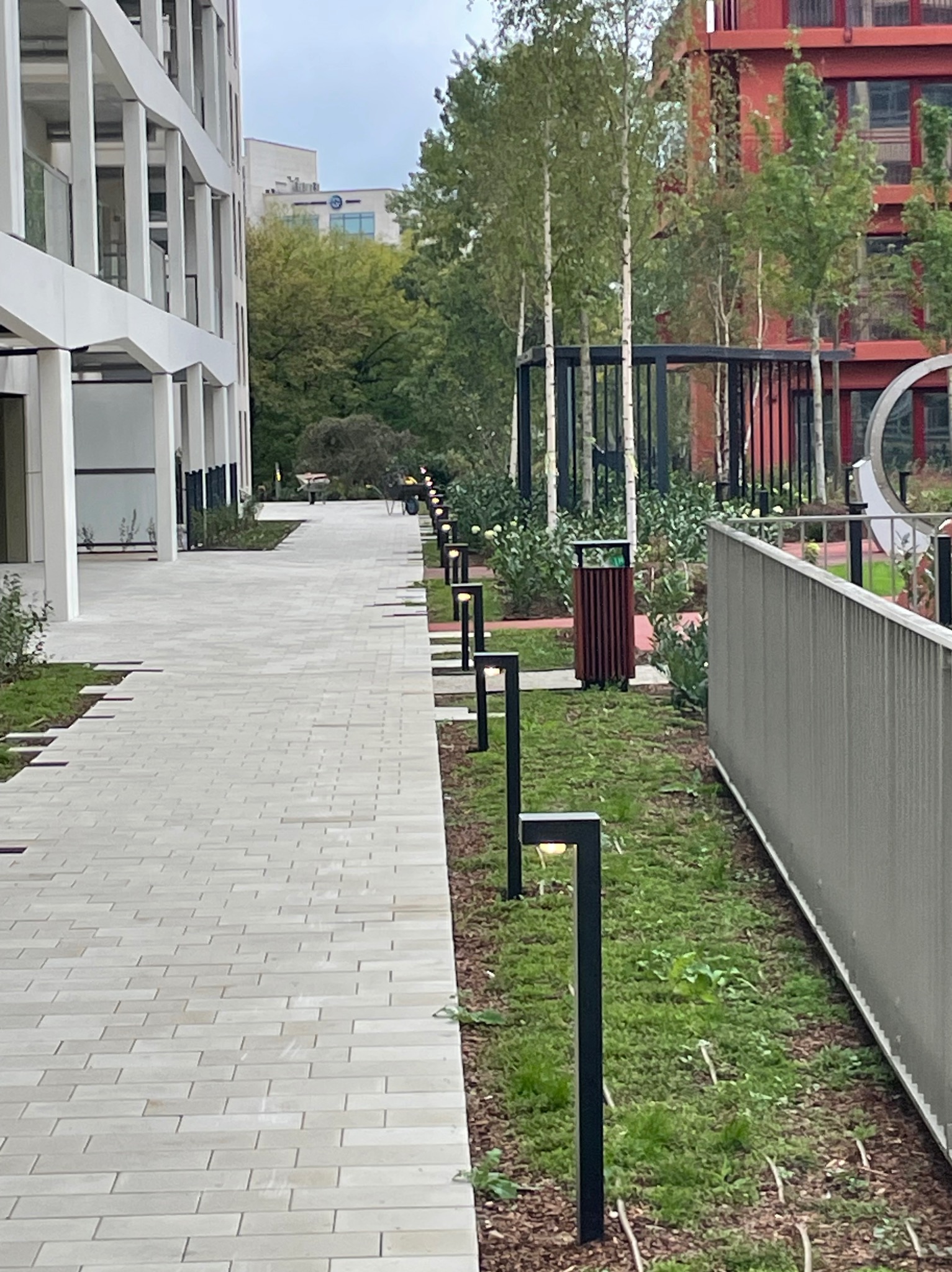 Ścieżka oświetlona lampami parkowymi przy nowoczesnym budynku. Lampy wzdłuż chodnika, trawnik, elementy małej architektury, drzewa w tle.