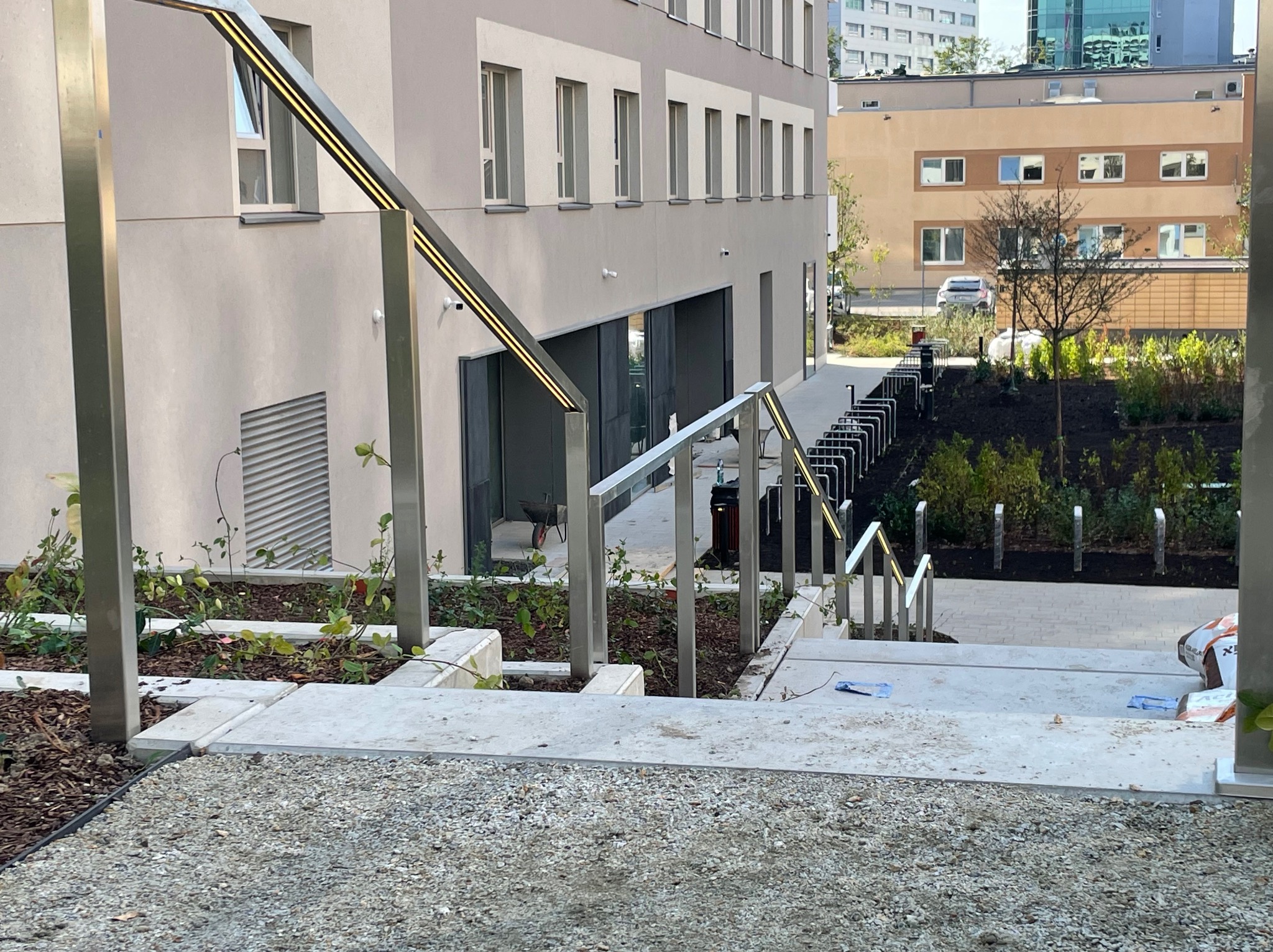 Nowoczesne oświetlenie schodów zewnętrznych z metalowymi barierkami i ledami, prowadzących do budynku w stylu minimalistycznym z widokiem na zieleń i parking.