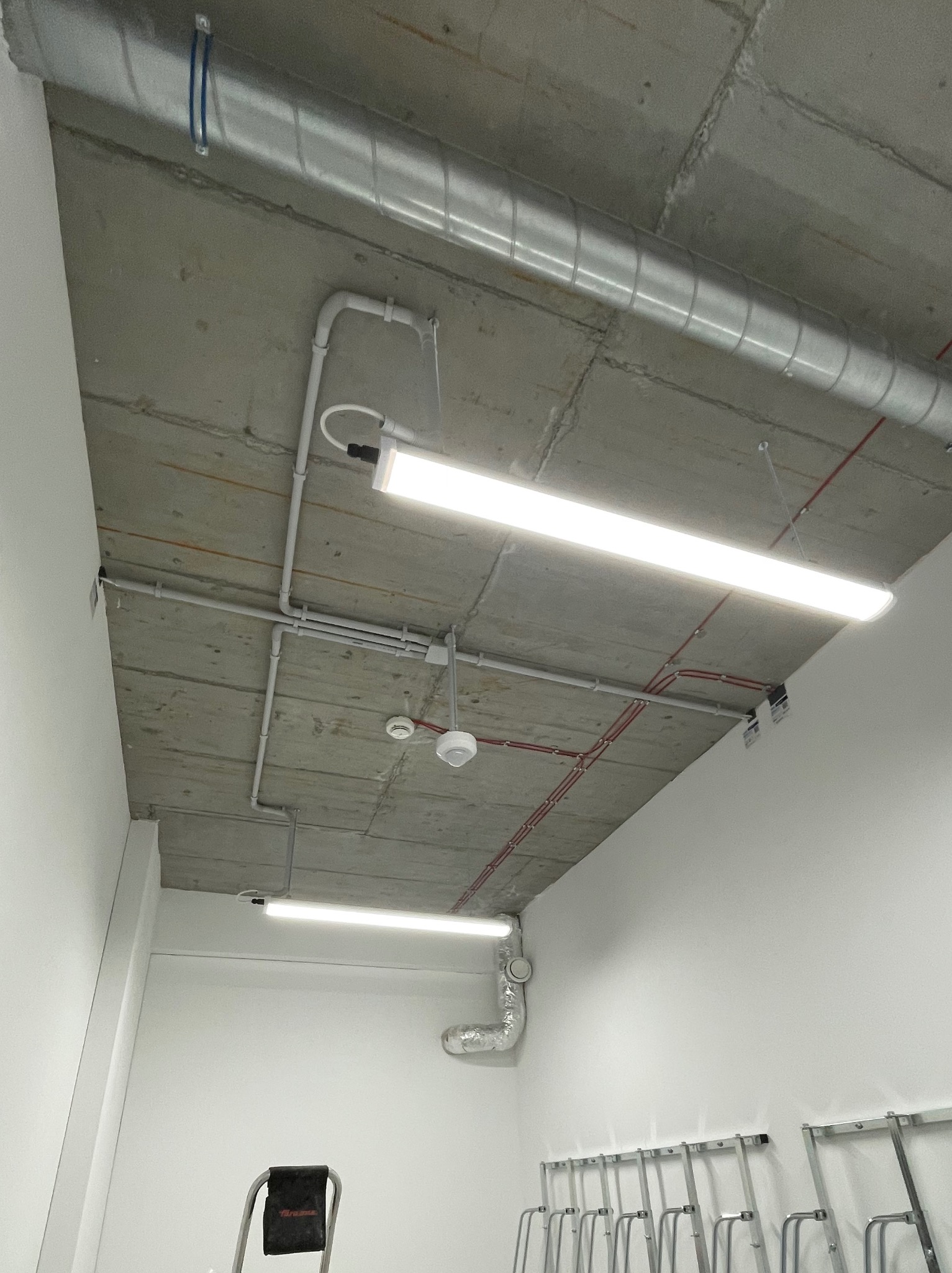 Zamontowane, podwieszone lampy liniowe LED na betonowym suficie, widoczne rury wentylacyjne i kable, drabina aluminiowa w rogu pomieszczenia.