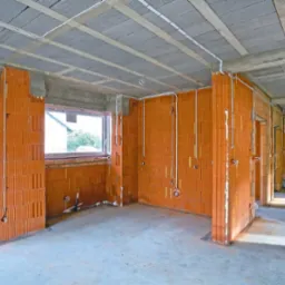 Wnętrze surowego budynku w Płońsku z widoczną instalacją elektryczną w peszlach na ceglanych ścianach, betonowy sufit z widocznymi elementami konstrukcyjnymi, okno z widokiem na budynek.