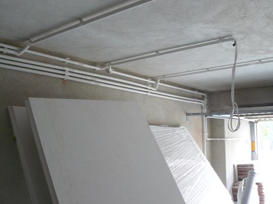 Białe rury elektryczne prowadzone pod sufitem w garażu, z widocznymi elementami mocującymi, oraz składowane płyty gipsowe i folia ochronna.