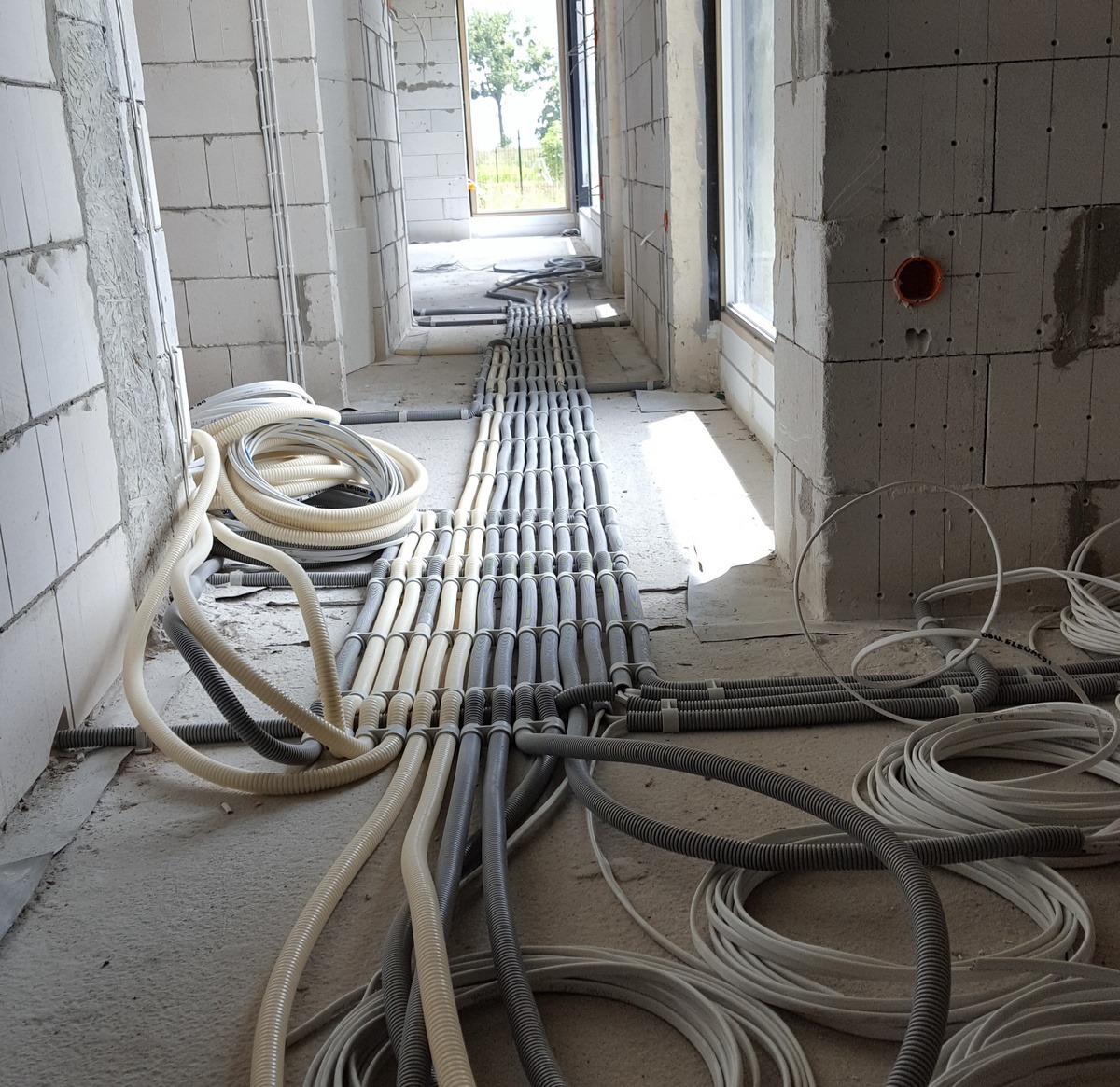 Wnętrze w stanie surowym z widocznymi peszlami układanymi w korytarzu, część ułożona równolegle, część w zwojach, na ścianie puszka instalacyjna.