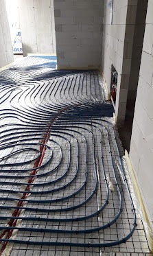 Rozłożone rury ogrzewania podłogowego na folii izolacyjnej, przymocowane do siatki zbrojeniowej w surowym wnętrzu budynku z bloczków.