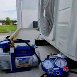 Zewnętrzna jednostka klimatyzacji Rotenso zamontowana na betonowym fundamencie, obok widoczna pompa próżniowa R32 i manometry serwisowe, w tle dom z szarą dachówką.