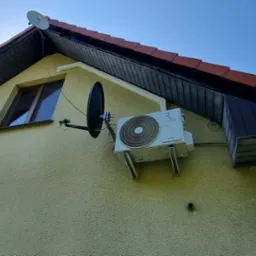 Zewnętrzna jednostka klimatyzacji Rotenso zamontowana na elewacji domu, obok okna i anteny satelitarnej, widok z dołu.