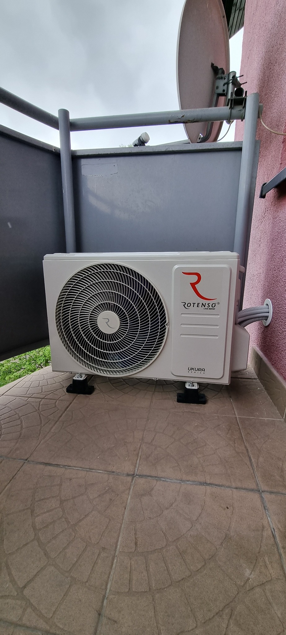Biała jednostka zewnętrzna klimatyzacji Rotenso Ukura zamontowana na balkonie, z widocznym przewodem w peszlu przechodzącym przez ścianę budynku.