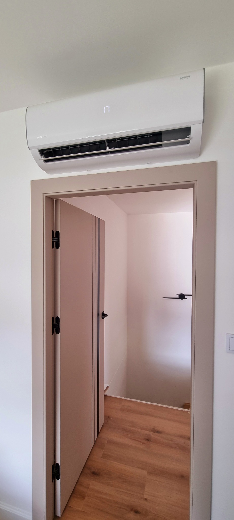 Biała jednostka klimatyzacji zamontowana nad drzwiami z widocznym wyświetlaczem temperatury, w tle widoczny korytarz z drewnianą podłogą.
