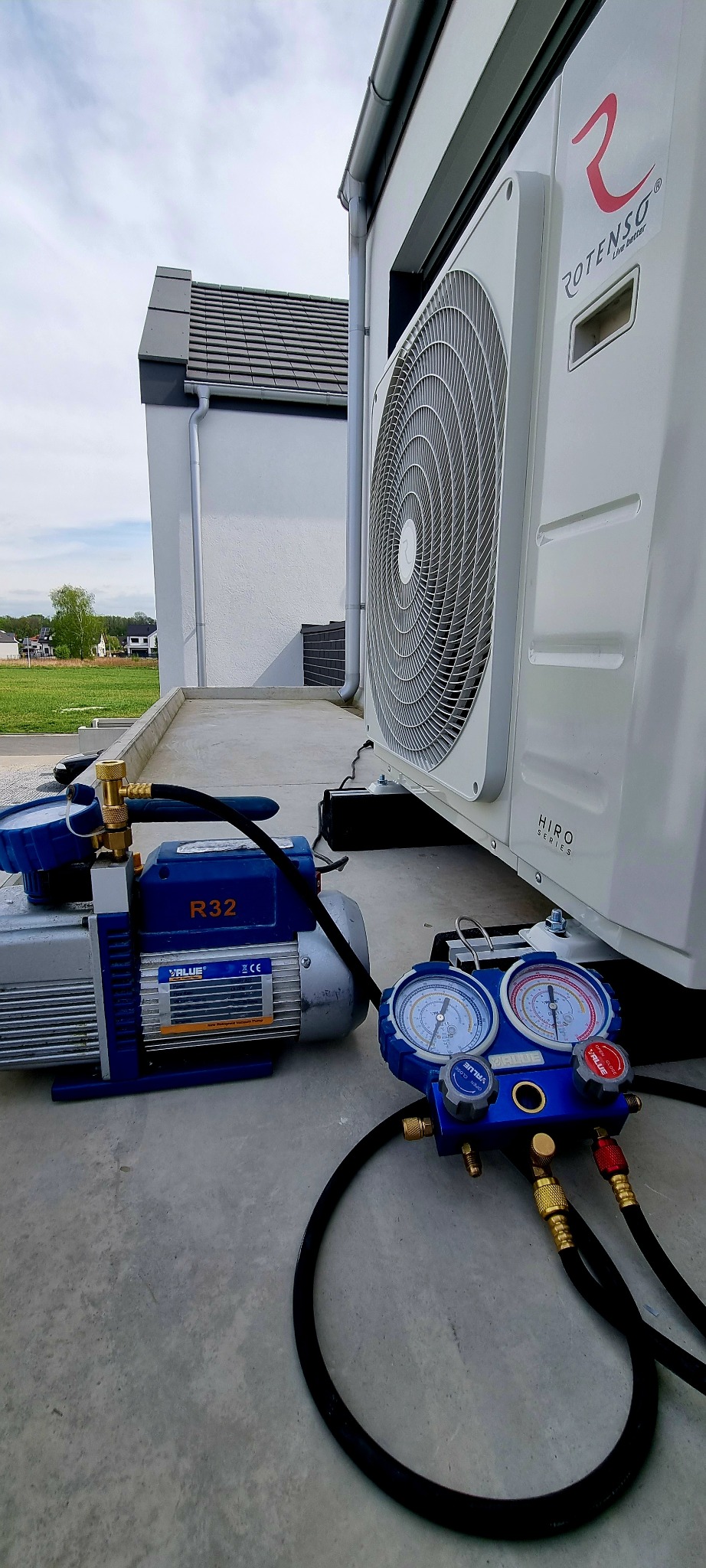 Zewnętrzna jednostka klimatyzacji Rotenso zamontowana na betonowym fundamencie, obok widoczna pompa próżniowa R32 i manometry serwisowe, w tle dom z szarą dachówką.