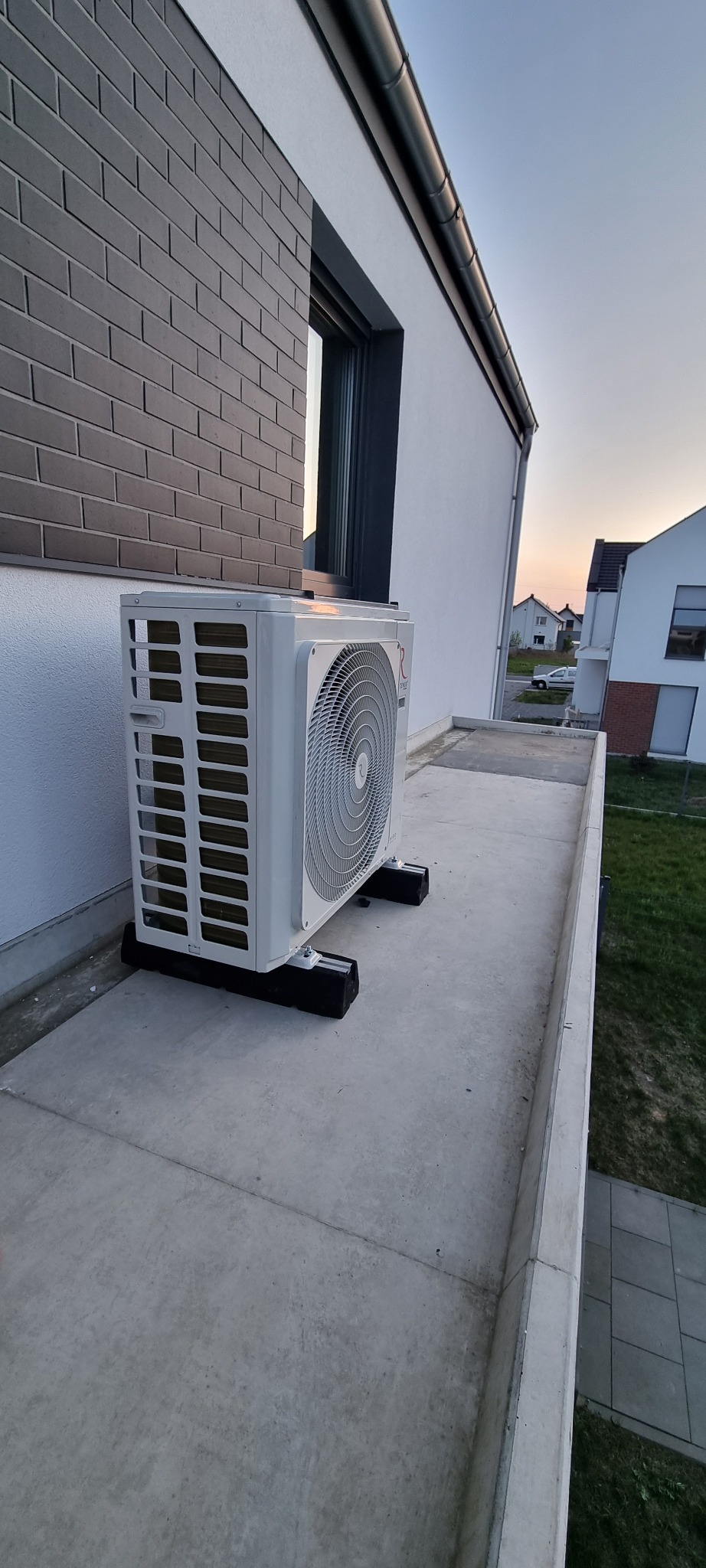 Zewnętrzna jednostka klimatyzacji zamontowana na płaskim dachu nowoczesnego domu jednorodzinnego, widok z góry pod lekkim kątem, w tle inne budynki i zachodzące słońce.