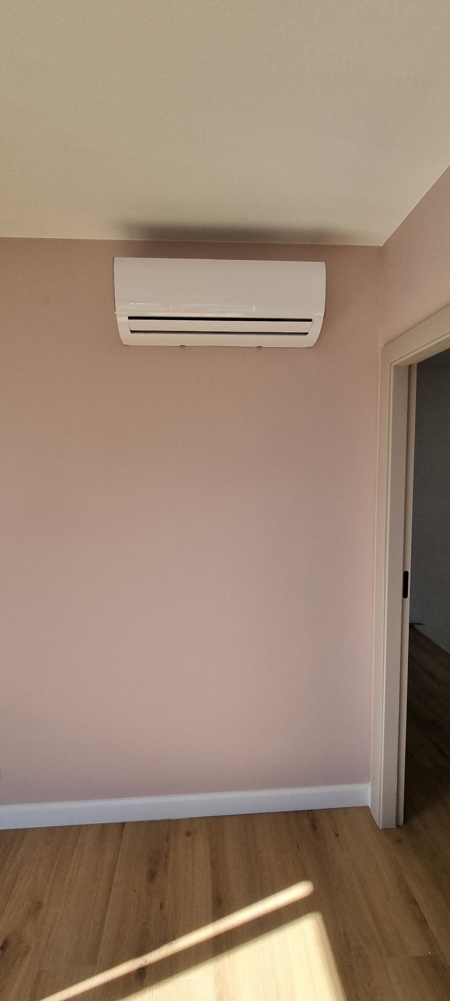 Biały klimatyzator ścienny zamontowany w pokoju z różowymi ścianami i drewnianą podłogą, widok z przodu, drzwi po prawej stronie.