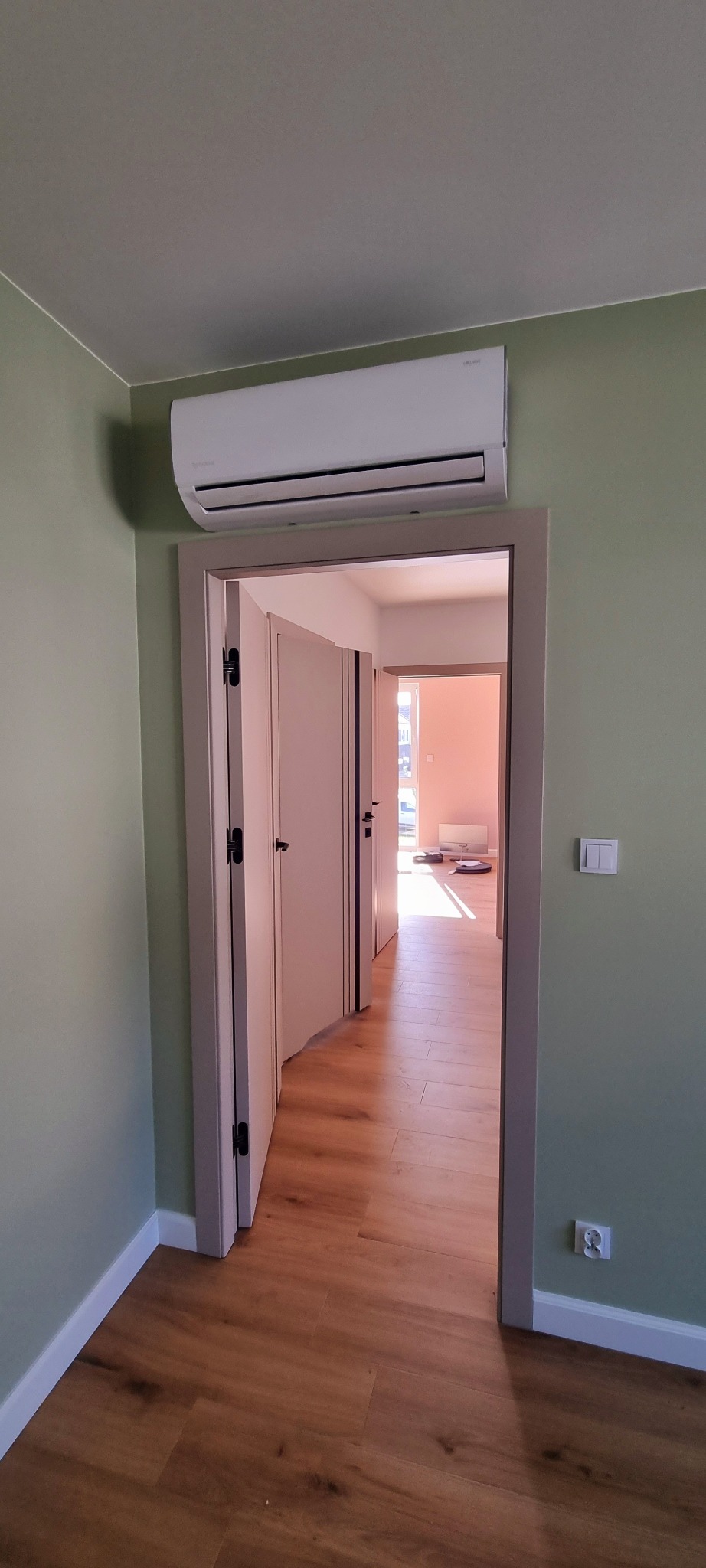 Biały klimatyzator ścienny zamontowany nad drzwiami w pokoju z zielonymi ścianami i drewnianą podłogą, widok na korytarz z otwartymi drzwiami i wpadającym światłem słonecznym.